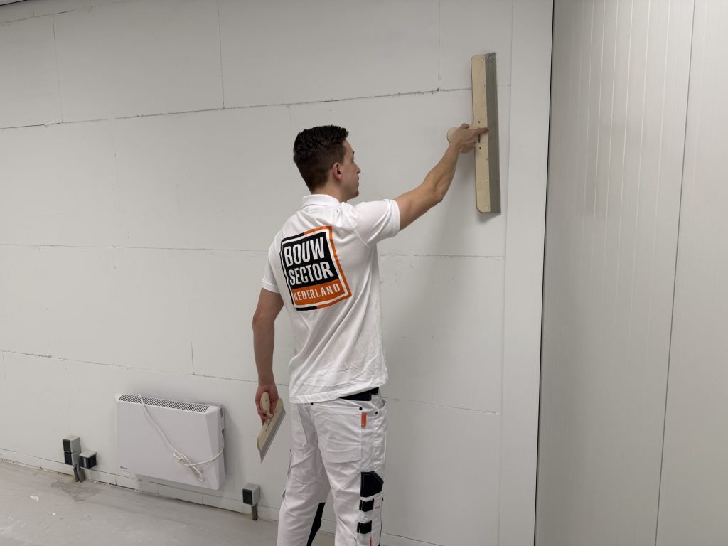 Stukadoor van Bouwsector Nederland zet een brede spaan tegen een wand met plaatnaden, naast een verwarmingspaneel