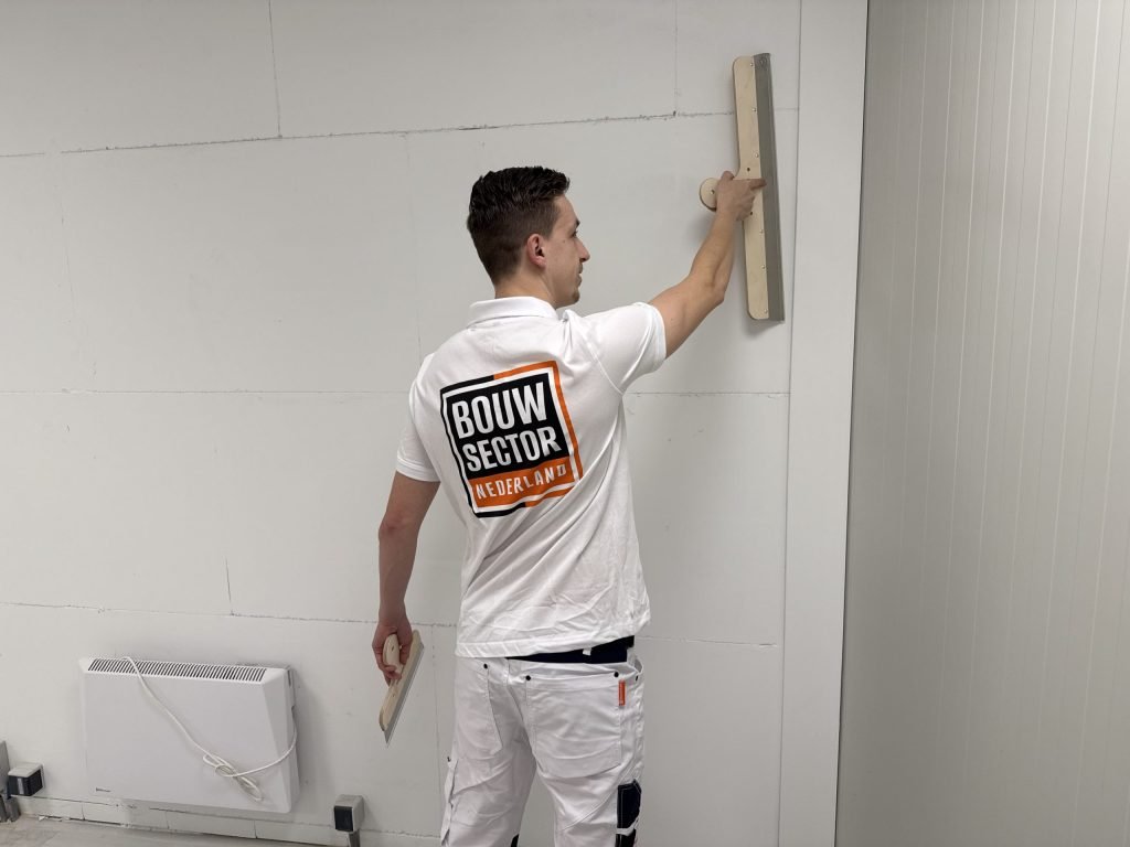 Stukadoor van Bouwsector Nederland zet een brede spaan tegen een wand met zichtbare plaatnaden